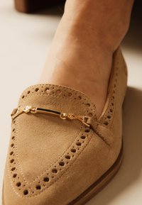 Κοντινό πλάνο ενός μοκασίνου από καφέ δέρμα σουέτ με διάτρητες λεπτομέρειες και χρυσή μεταλλική διακοσμητική μπάρα στην κορυφή, φορεμένο σε πόδι.