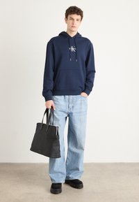 Felpa navy con tasca a marsupio e lacci argento, abbinata a jeans azzurri larghi e una borsa tote nera, tenuta nella mano destra.