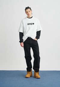 Maglietta grafica bianca indossata sopra una camicia a maniche lunghe nera, abbinata a jeans neri e stivali color cuoio; presenta una stampa sulla maglietta.