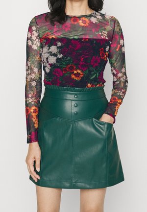 Femme portant un haut transparent à manches longues fleuri et coloré, associé à une mini-jupe en cuir verte taille haute avec des boutons et des poches.