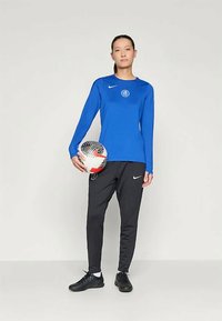 Sininen pitkähihaine urheilupaita logolla, mustat urheiluhousut ja mustat lenkkarit. Pitelee mustavalkoista jalkapalloa.