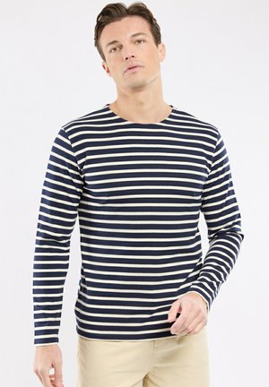 Homme portant un t-shirt à manches longues rayé horizontalement bleu marine et blanc et un pantalon beige sur un fond blanc uni.