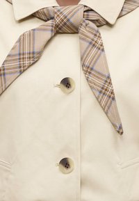 Manteau beige boutonné avec une écharpe à carreaux marron et bleue nouée autour du col, montrant deux grands boutons ronds et une partie de deux poches.