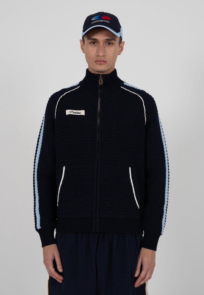 Veste zippée marine avec un motif en tricot texturé, accents bleu clair et blancs rayés le long des manches, et poches avant. Comporte un patch logo sur la poitrine.