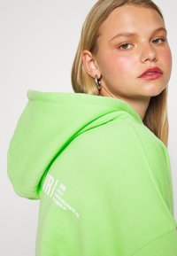 Sweatshirt verde néon com capuz, apresentando texto branco do lado esquerdo. Tecido suave com um corte solto e detalhes de costura visíveis.