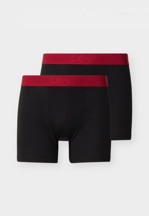 Jeune homme debout, les mains derrière le dos, portant un boxer noir avec une ceinture rouge, sur un fond blanc uni.