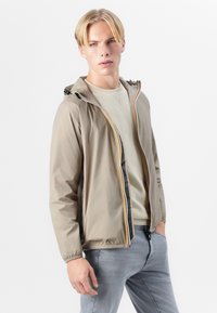Chaqueta ligera color beige con capucha, puños elásticos y un cierre frontal con ribetes multicolores, llevada sobre un suéter gris claro y jeans.