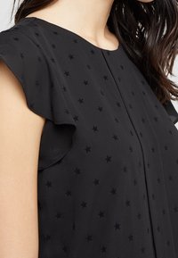 Svart blus med stjärnmönster, med fladdrande ärmar, mjuk textur och avslappnad passform. Detaljstygn synligt längs halsringningen.