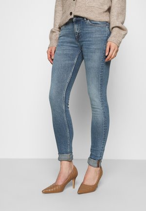 Mujer con jeans ajustados azul claro con puños enrollados, cárdigan de punto beige y zapatos de tacón alto color cuero con punta puntiaguda, de pie frente a un fondo liso.