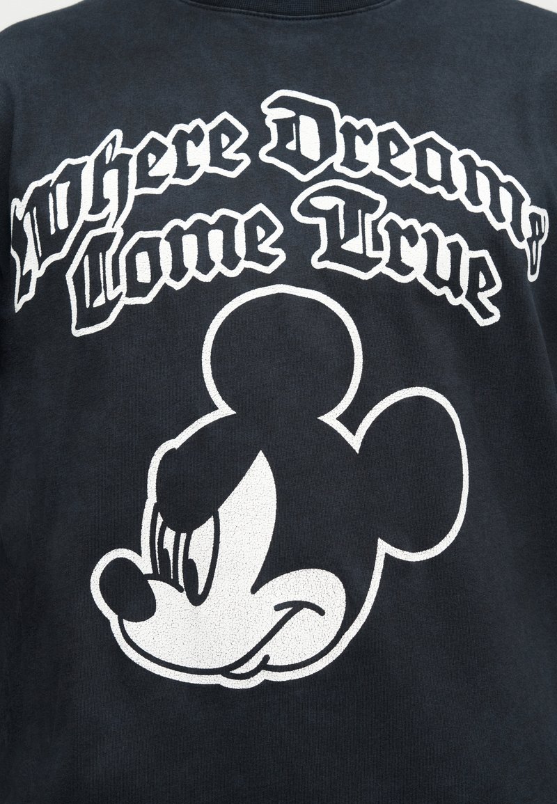 Sort skjorte med hvid tekst "Hvor drømme går i opfyldelse" over en hvid kontur af Mickey Mouses smilende ansigt.
