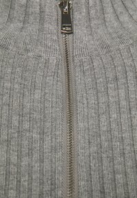Grauer gerippter Strickpullover mit einem auffälligen silbernen Reißverschluss in der Mitte, der strukturierte vertikale Linien und einen weichen Stoff aufweist.
