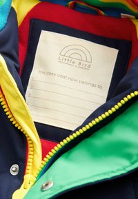Večbarvna notranjost plašča s rumeno, zeleno in mornarsko modro blago, ki vključuje oznako za personalizacijo ter mehak rdeč podstavek.