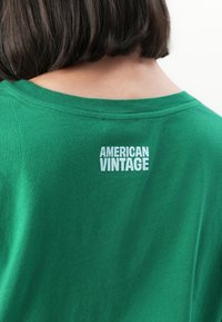 Asmuo su trumpais tamsiais plaukais, vilkintis žalią marškinį, ant nugaros viršuje yra spaudinys "AMERICAN VINTAGE" baltos spalvos.