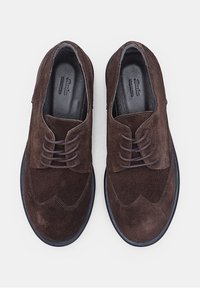 Tumši brūni zamša lace-up apavi ar melniem zolemi, skatīti no augšas uz gaiša pelēka fona.