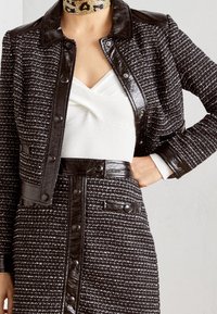 Veste en tweed noir et blanc avec des accents en cuir noir, présentant un col et des boutons-pression. Haut blanc avec un motif texturé en dessous.