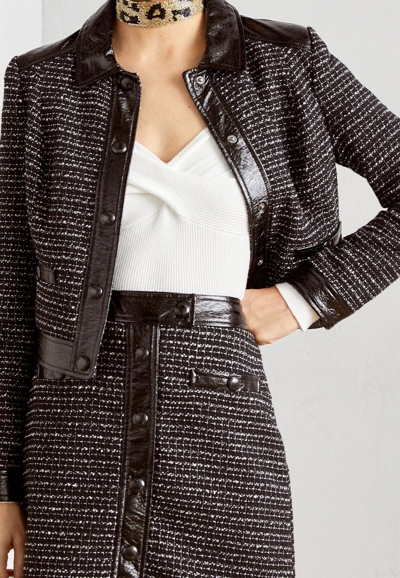 Veste en tweed noir et blanc avec des accents en cuir noir, présentant un col et des boutons-pression. Haut blanc avec un motif texturé en dessous.
