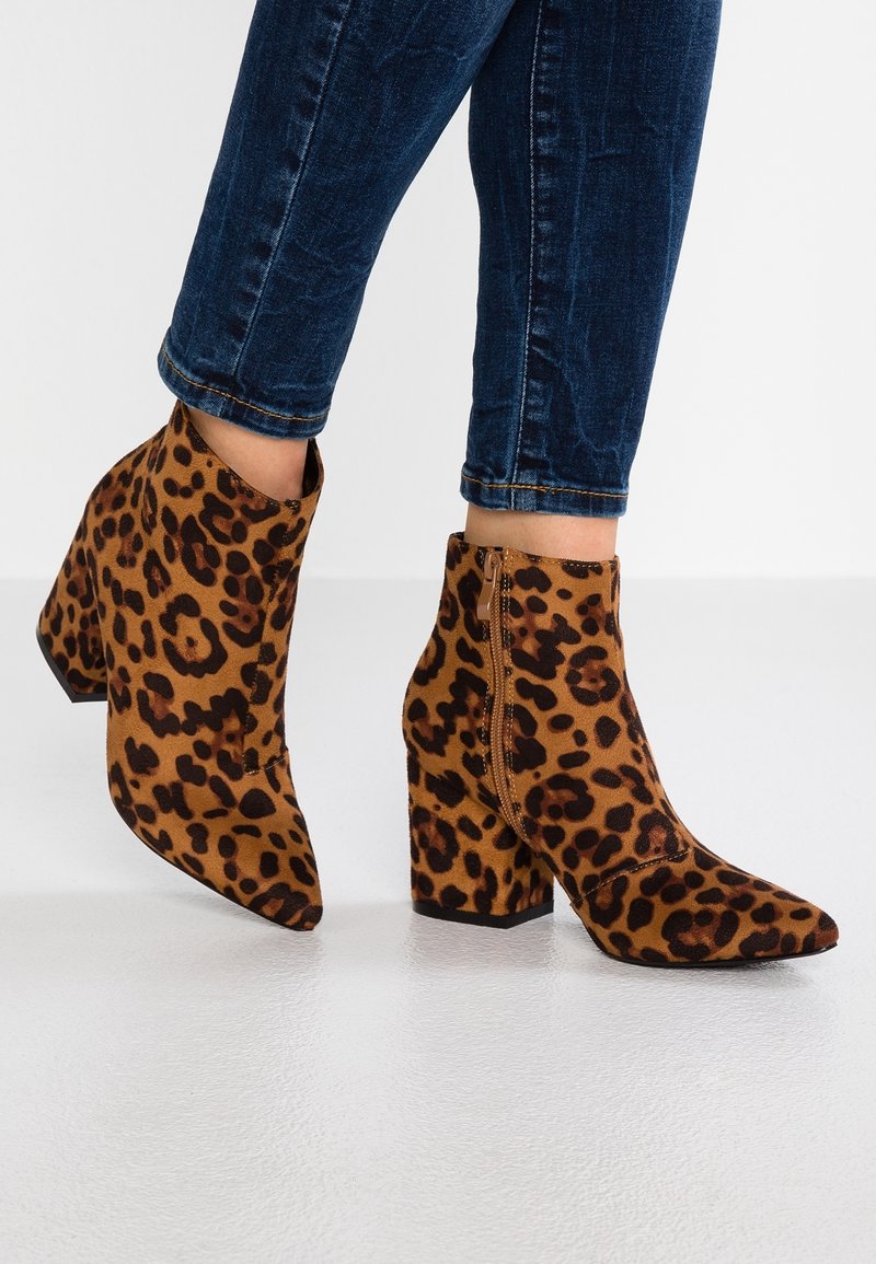 Personne portant des bottines à talons haut léopard montant à la cheville, associées à un jean skinny bleu foncé retroussé sur un sol blanc.