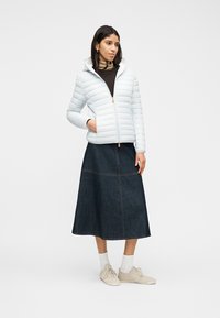 Γυναίκα που φοράει λευκό πουπουλένιο μπουφάν με κουκούλα, μαύρο τοπ, σκούρα τζιν μίντι φούστα, λευκές κάλτσες και μπεζ αθλητικά παπούτσια, στέκεται με το ένα χέρι στην τσέπη.