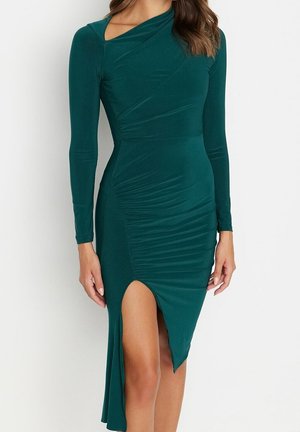 Kobieta ubrana w dopasowaną, ciemnozieloną sukienkę z długim rękawem, z asymetrycznymi marszczeniami i rozcięciem na lewej stronie sięgającym uda.