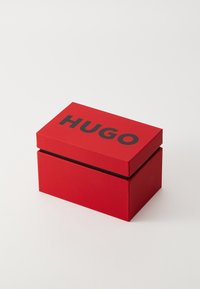 Rode rechthoekige doos van karton met een gladde textuur, met bovenop het woord "HUGO" in grote zwarte letters.