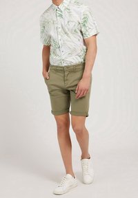 Camisa de botones de manga corta con un patrón de hojas verdes, combinada con pantalones cortos caqui enrollados. Zapatillas blancas completan el conjunto.