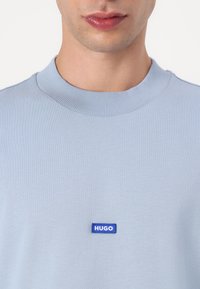 Svijetloplavi pulover s okruglim izrezom, napravljen od mekane tkanine, s rebrastim ovratnikom i malim pravokutnim plavim logom "HUGO" na prsima.