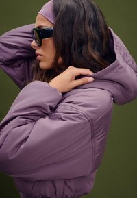 Helllila gefütterte Jacke mit Kapuze und gestepptem Design, die eine Nahaufnahme des Ärmels und des Kragens zeigt. Mattes Finish und glattes Material.