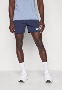 Calções de desporto azuis com texto branco "BORN", combinados com meias brancas e sapatilhas. O tecido parece leve e respirável.