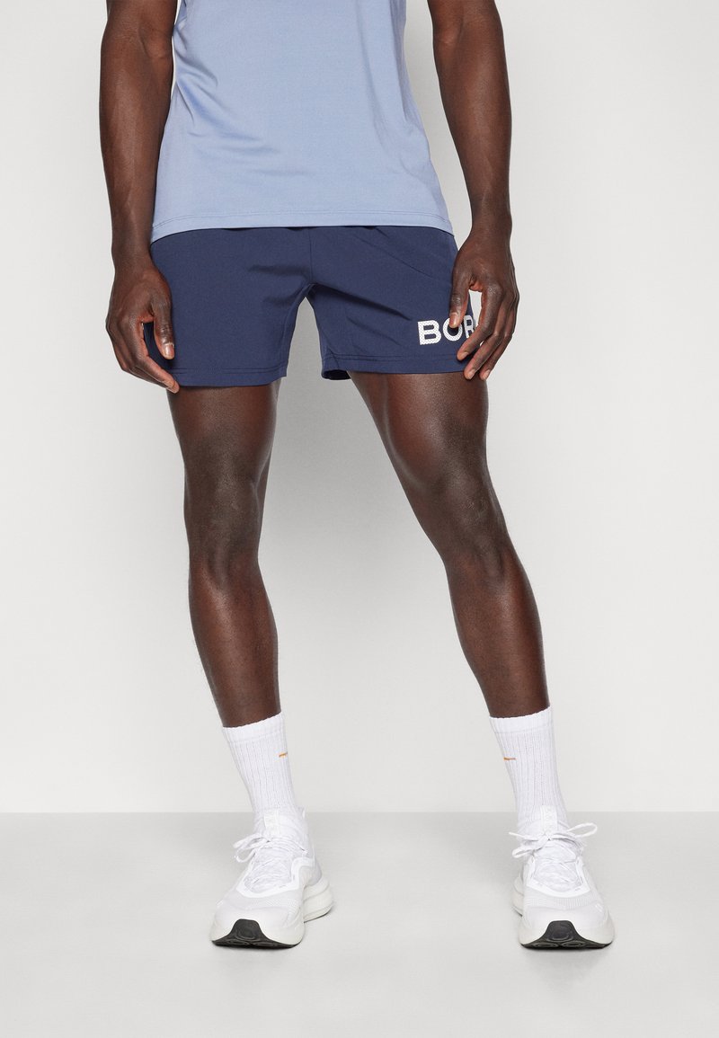 Calções de desporto azuis com texto branco "BORN", combinados com meias brancas e sapatilhas. O tecido parece leve e respirável.