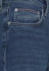 Vaqueros de mezclilla azul oscuro con un diseño de bolsillo clásico que incluye dos remaches plateados y un acento de costura roja en el borde del bolsillo.