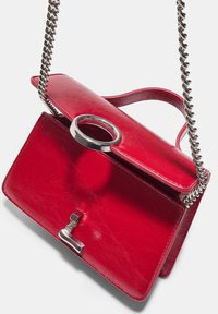 Red leather handbag with a textured finish, featuring a circular handle, silver chain strap, and a metal clasp. Compact design with sleek lines.