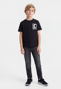 Vaaleahiuksinen poika seisoo musta Kani -t-paita päällään, kulutetut mustat farkut ja mustat lenkkarit yllään yksiväristä vaaleaa taustaa vasten.