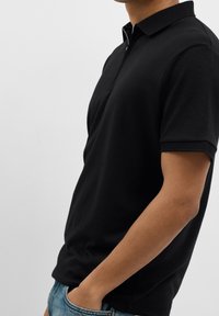 Svart polo med dragkedja vid kragen, kortärmad med strukturerad kant och lättviktigt tyg. Kombinerad med blå denimjeans.