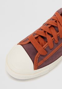 Purple suede sneakers with a cream rubber toe cap, orange laces, and contrasting brown accents on the sides and stitching details.