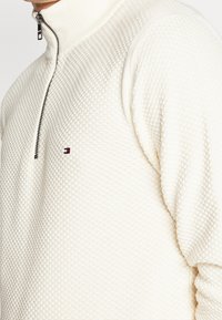 Detailní záběr na osobu v texturovaném krémovém pleteném svetru s čtvrtinovým zipem a malým červeným, bílým a černým logem na hrudi.