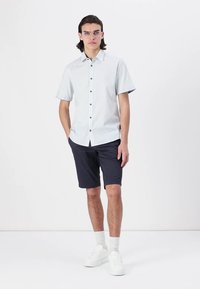 Camisa de botão de manga curta, azul claro, com botões pretos, combinada com calções escuros azul-marinho e sapatilhas brancas, apresentando meias brancas caneladas.