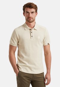 Beige Poloshirt mit kurzen Ärmeln, Knopfleiste und kleiner Brusttasche. Aus strukturiertem Stoff gefertigt, mit dunklen Knöpfen und aufgerollten Ärmeln.