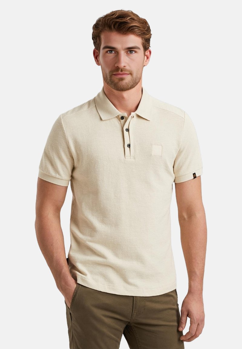 Beige Poloshirt mit kurzen Ärmeln, Knopfleiste und kleiner Brusttasche. Aus strukturiertem Stoff gefertigt, mit dunklen Knöpfen und aufgerollten Ärmeln.