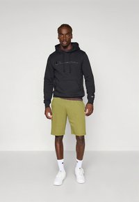 Camisola com capuz preta com o logótipo "Champion" bordado, calções verdes-oliva e ténis brancos. As meias são brancas com detalhes vermelhos. Roupa simples e casual.