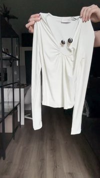 Langärmliges weißes Oberteil mit V-Ausschnitt, gerafftem Design an der Vorderseite, dekoriert mit zwei schwarzen Knöpfen und einer glatten Stoffstruktur.
