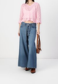 Top corto de punto rosa con escote en V y patrón de calados. Pantalones vaqueros de pierna ancha en azul claro con cintura ajustable. Bolso marrón con correa ajustable.