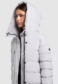 Šviesiai pilka pūkinė striukė su dideliu gobtuvu, priekiu su užtrauktuku, spaustukais ir raukšlėta tekstūra. Šoninės kišenės su skląsčių uždarymu.