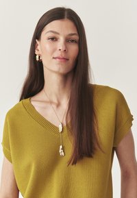 Femme avec de longs cheveux bruns portant des boucles d'oreilles en or, un collier pendentif en or et un haut en tricot à encolure en V jaune moutarde devant un fond uni.