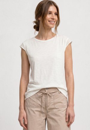 Kvinde med lysebrunt hår i en lav knold iført en hvid kortærmet top og beige bukser med forlommer og knyttet talje, stående mod en ensfarvet baggrund.