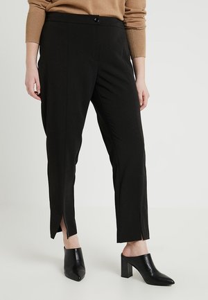 Femme portant un pantalon noir longueur cheville avec des fentes à l'avant, un haut marron à manches longues et des mules noires à talons sur fond blanc.