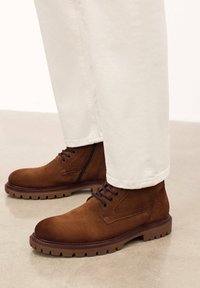 Stivaletti in suede marrone con punte rotonde, lacci e suole in gomma testurizzata. Abbinati a pantaloni chiari con risvolto.