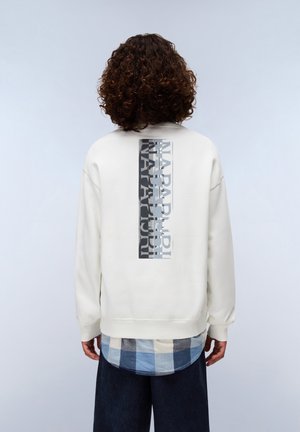 Sweat-shirt couleur crème avec un design graphique rectangulaire en gris et noir dans le dos. Superposé à une chemise à carreaux bleue et un pantalon foncé.