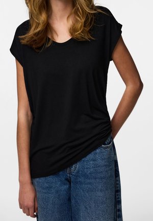 Femme aux cheveux ondulés mi-longs portant un haut noir à manches courtes et un jean en denim bleu, une main dans la poche.