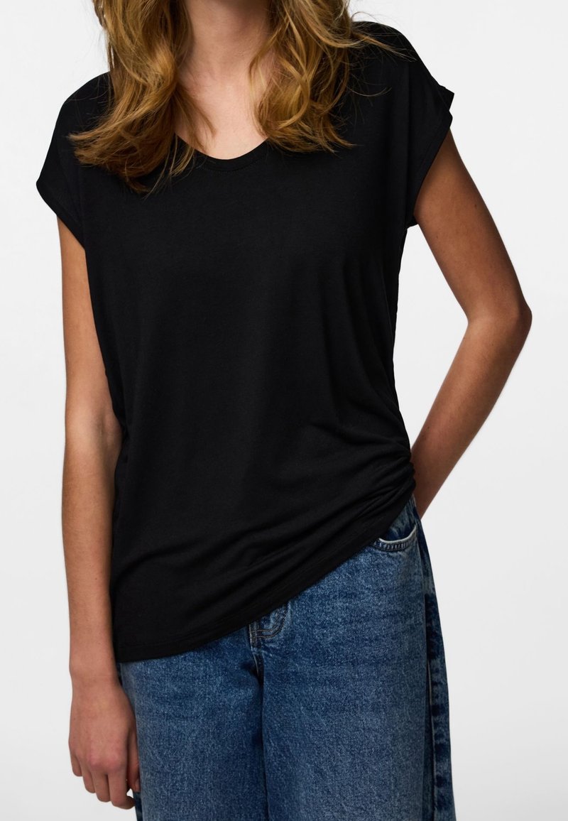 Kobieta o falowanych włosach do ramion, ubrana w czarny top z krótkim rękawem i niebieskie dżinsy, z jedną ręką w kieszeni.