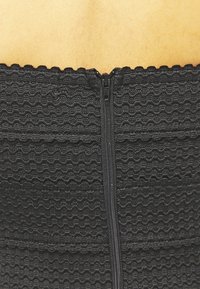 Vêtement noir texturé avec des motifs ondulés, doté d'une fermeture éclair centrale. Le tissu semble élastique et ajusté. Vue rapprochée du bord supérieur.